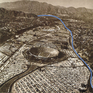 A black-and-white aerial view of Rose Bowl from 1962 surrounded by parking lot full of vehicles. A blue line has been drawn over the image to illustrate an alternate route for the Foothill Freeway/I-210 section that would have run adjacent to the venue's parking lot.