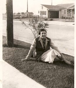 Black woman in a jacket and dress sitting in front of a small tree in between a sidewalk and a street, with a house and a "Model Homes" sign on the other side of the street behind