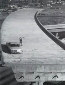 Councilmember Joe Colla, posing with arms outstretched, on an incomplete freeway ramp bridge, next to a car